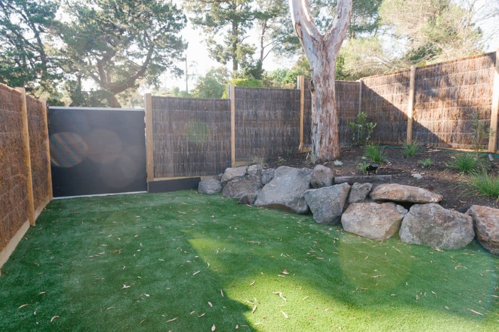 backyard lawn around a boulder retaining wall with natural trees and wicker fence